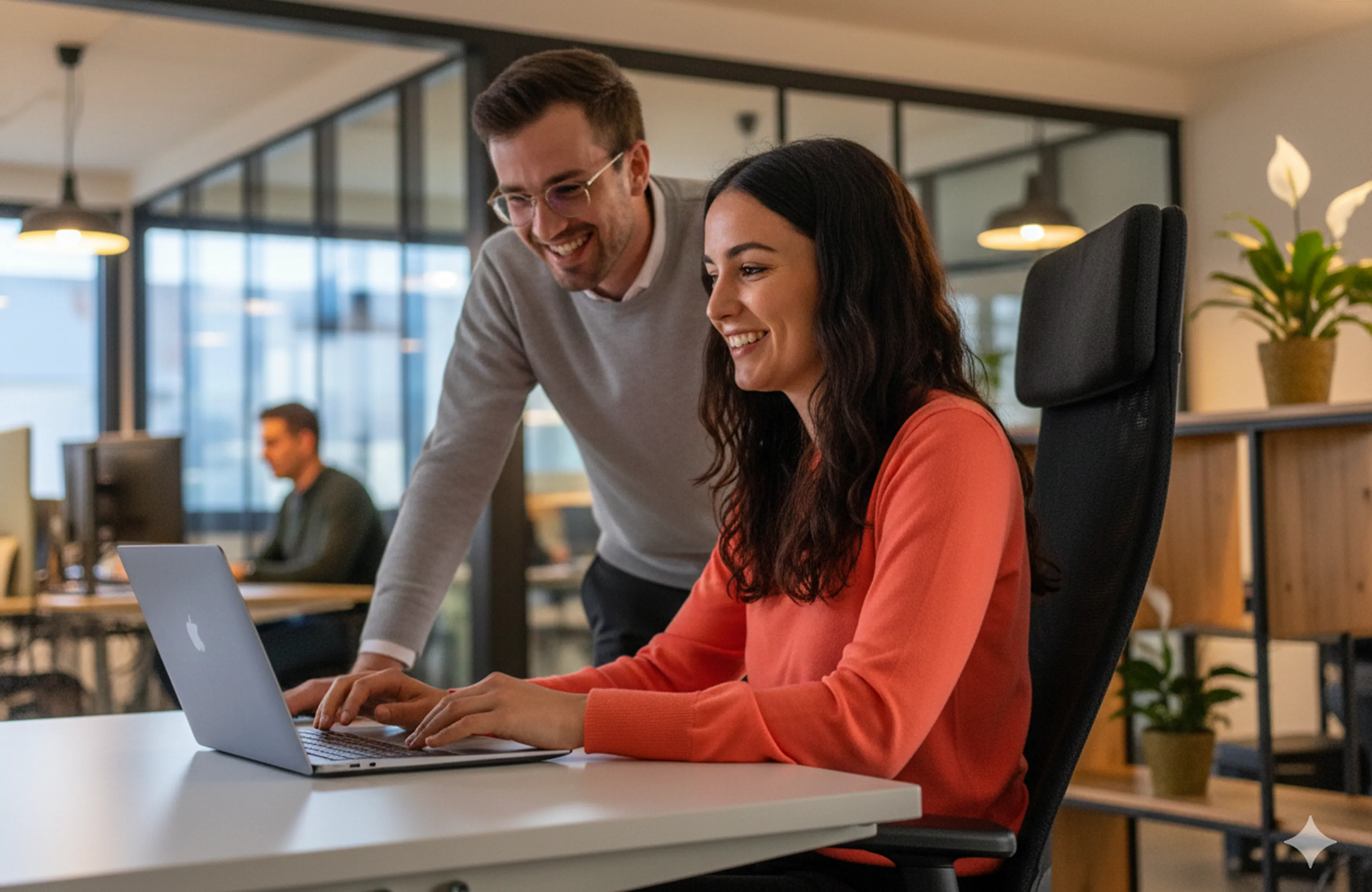 Two smiling Positive group employees collaborating on a laptop in a modern office environment.