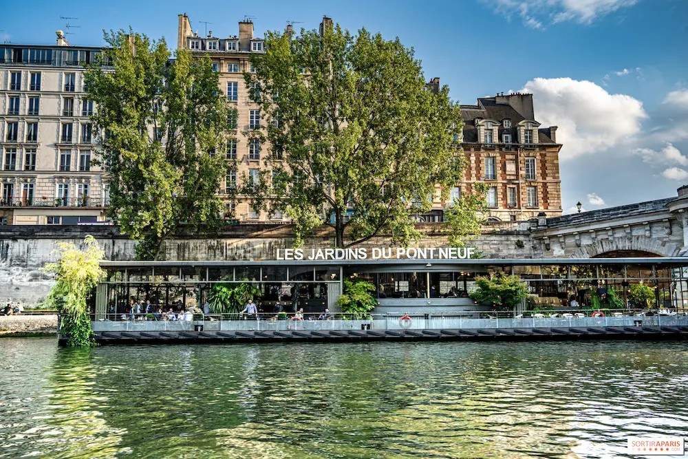 Floating restaurant 'Les Jardins du Pont Neuf' on a river with green trees and historic buildings in the background under a blue sky.